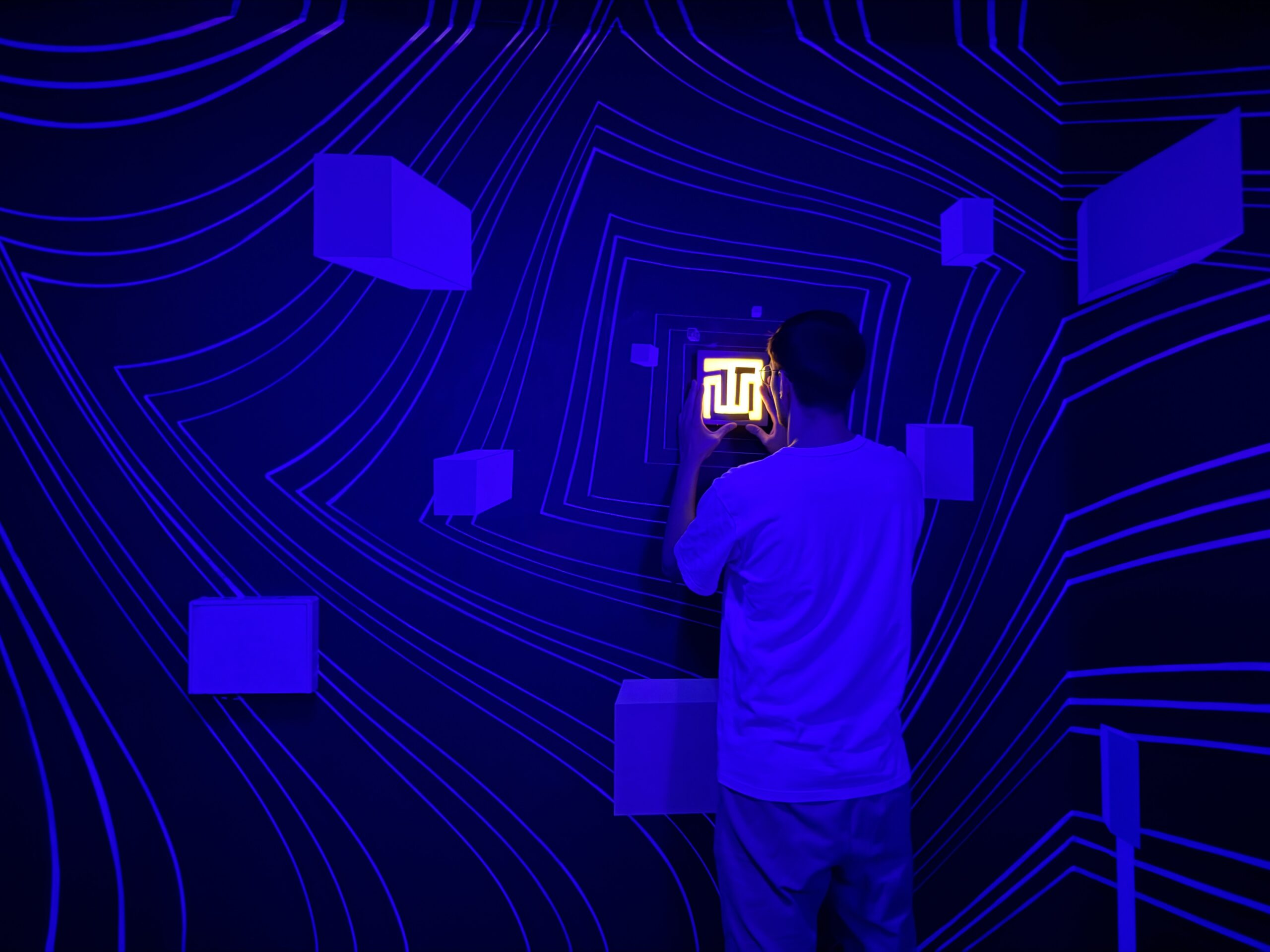 Dans The Bunker, une personne vue de dos, dans une salle aux murs bleus peints de lignes lumineuses, tient un petit carré éclairé contre le mur