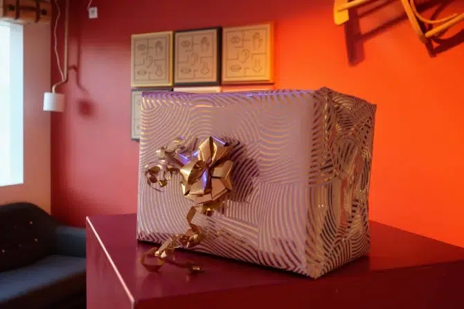 Un paquet cadeau orné d’un nœud doré sur le côté, posé sur une table rouge dans le décor orange de Tactisens
